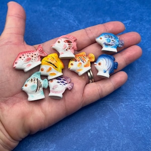 May include: A collection of small, ceramic fish figurines in various colors, including red, yellow, blue, and purple. Each fish has unique patterns and details, with white accents. The figurines are held in a hand against a blue background.