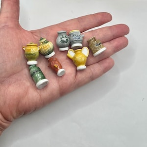 May include: A collection of eight miniature ceramic jars and pitchers in various colors including yellow, green, grey, brown, and orange. Each small vessel features a unique shape, a white base, and handwritten text labels on the exterior surface.
