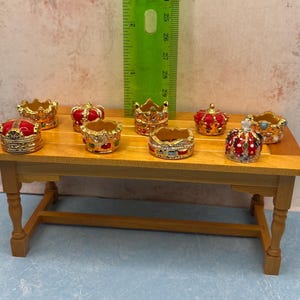 May include: A collection of miniature decorative crowns in various colors, including gold, red, and blue, are displayed on a small wooden table. The crowns have intricate details and are likely made of metal or a similar material. A green ruler stands behind the crowns.