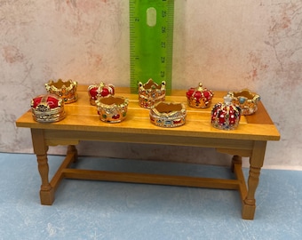 May include: A collection of miniature decorative crowns in various colors, including gold, red, and blue, are displayed on a small wooden table. The crowns have intricate details and are likely made of metal or a similar material. A green ruler stands behind the crowns.