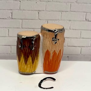 May include: A miniature ceramic conga drum set with a light tan drumhead and a silver rim. The drums are painted with shades of brown, orange, and yellow. The set is placed on a white surface against a white brick wall.