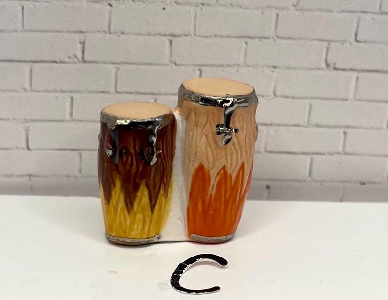 May include: A miniature ceramic conga drum set with a light tan drumhead and a silver rim. The drums are painted with shades of brown, orange, and yellow. The set is placed on a white surface against a white brick wall.