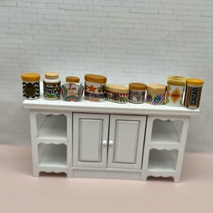 May include: A collection of miniature ceramic jars with lids, arranged on a white cabinet. The jars feature various designs and labels, including the word "THE" and "CACAO". The cabinet has two doors and open shelves.