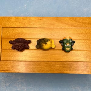 May include: A wooden box with three decorative face carvings. The carvings are brown, yellow, and green, each with unique features. The box is made of light-colored wood and sits on a blue surface.