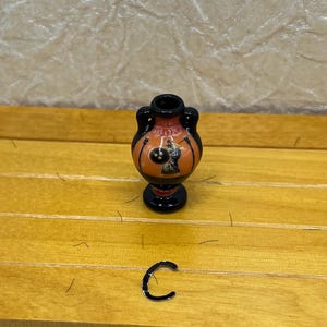 May include: A small, decorative vase with a black base and handles, featuring an orange body with a black and white illustration. The vase is set on a wooden surface. A small, black, C-shaped object is also present.