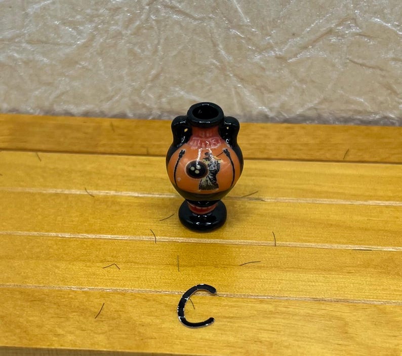 May include: A small, decorative vase with a black base and handles, featuring an orange body with a black and white illustration. The vase is set on a wooden surface. A small, black, C-shaped object is also present.
