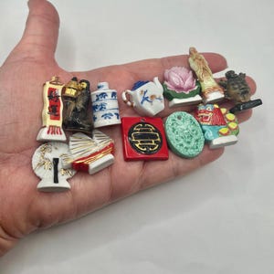 May include: A collection of small, colorful ceramic figurines, including a teapot, a lotus flower, and various Asian-inspired designs. The figurines are held in a hand, showcasing their intricate details and vibrant colors. The collection includes red, white, blue, green, and gold accents.
