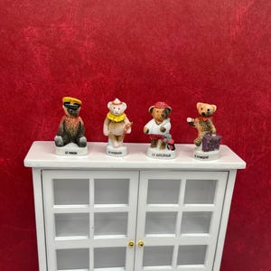 May include: Four small, decorative bear figurines on a white cabinet. Each bear is dressed in a different outfit, including a sailor, a musician, a golfer, and a traveler. The figurines are set against a red background.