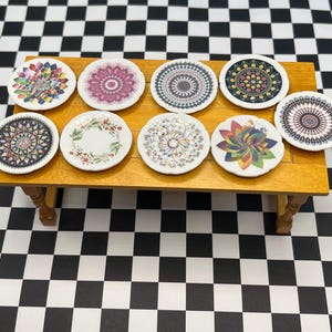 May include: A collection of eight small, decorative ceramic plates with various colorful mandala and floral designs. The plates are arranged on a miniature wooden table, set against a black and white checkered background.