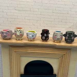 May include: A collection of six small, ceramic face planters with various expressions. Each planter is painted with different colors and facial features, including winking eyes, surprised expressions, and closed eyes. The planters are displayed on a wooden shelf.