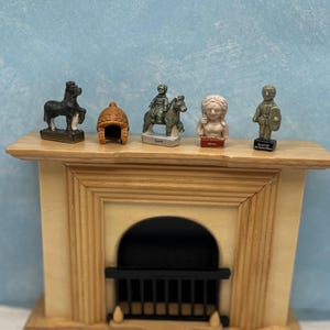 May include: Miniature figurines of historical figures and objects, including a horse, helmet, rider on a horse, bust, and a figure with a shield, are displayed on a small wooden mantelpiece. The figurines are various colors and sizes.