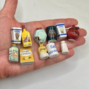 May include: A collection of eleven miniature ceramic kitchen canisters and containers featuring various shapes and colors. The items include labels such as Farine, Rhum, Cafe, Sel, Pates, Sucre Vanille, and Riz, with painted details like wheat, coffee beans, and vanilla flowers.