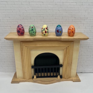 May include: A miniature wooden fireplace with a light wood finish, featuring a black grate and arched opening. Five decorative, colorful Easter eggs with floral and heart designs rest on the mantelpiece. The eggs are pink, green, yellow, blue, and orange.