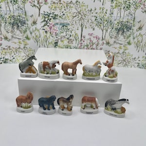 May include: A collection of ten ceramic horse figurines, each with a unique design and color scheme. The figurines are displayed on a white surface, showcasing various breeds like Appaloosa and Paint Horse. The background features a floral wallpaper.