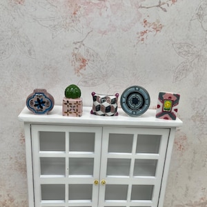 May include: Miniature ceramic decorative items, including a vase, a cactus, a pillow, a circular object, and a square object, are displayed on a white cabinet. The items feature various colors and patterns, such as blue, green, pink, and geometric designs.