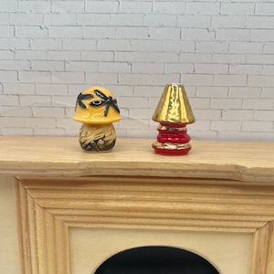 May include: Two miniature ceramic lamps. One is shaped like a mushroom with a yellow cap and black accents. The other is a red and gold lamp with a conical shade. Both are displayed on a wooden surface against a white brick wall.