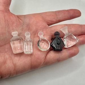 May include: Five miniature perfume bottles in various shapes and designs. The bottles are clear, frosted, and black, with different stopper styles. The bottles are held in a hand against a white background.