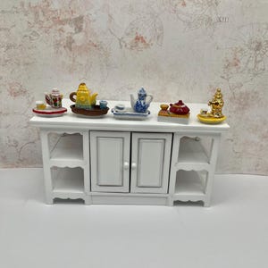 May include: A white cabinet displays miniature tea sets. The sets feature colorful teapots, cups, and saucers in various designs, including floral and gold accents. The cabinet has two doors and open shelves.