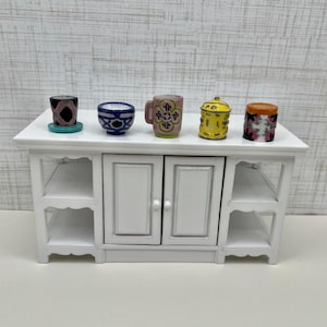 May include: A miniature white cabinet with a countertop displaying five small, decorative ceramic items. These include a small black and pink patterned cup, a blue and white bowl, a mug with a floral design, a yellow jar, and a small jar with a pink and black pattern.