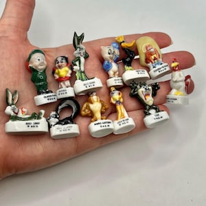 May include: A collection of twelve miniature ceramic Looney Tunes character figurines, each mounted on a small white base with the character name and copyright text printed in black. The set includes Bugs Bunny, Elmer Fudd, Petunia Pig, Porky Pig, Daffy Duck, Yosemite Sam, Foghorn Leghorn, Pepe Le Pew, Marc Antony, Lola Bunny, and Taz.