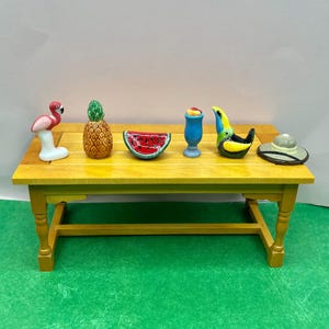 May include: A yellow miniature table displays several ceramic figurines. These include a pink flamingo, a pineapple, a watermelon slice, a blue cocktail, a toucan, and a sun hat. The table is set on a green surface.