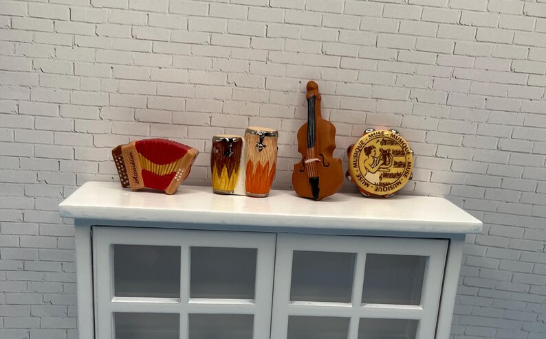 May include: A collection of miniature musical instruments, including an accordion, drums, a violin, and a tambourine. The accordion is red and gold, the drums are brown and orange, the violin is brown, and the tambourine has a beige face with text.