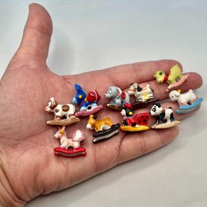 May include: A collection of miniature rocking horse figurines in various animal designs, including a cow, elephant, and panda. Each figurine is brightly colored and rests on a small, curved base. The figurines are small, likely under 2 inches in length.