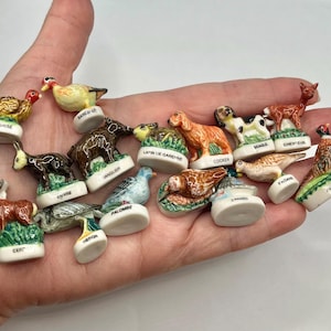 May include: A collection of small, hand-painted ceramic animal figurines, each mounted on a white base with a printed label. The set includes various wildlife such as a deer, pheasant, rabbit, boar, and dog, all featuring detailed, colorful glazes.