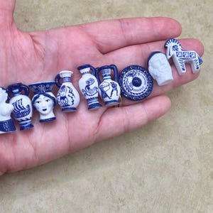 May include: A collection of nine miniature ceramic figurines featuring white surfaces with blue painted details. The set includes various shapes such as a bust, amphorae, a circular plate, a head, and a horse, all decorated with classical motifs.