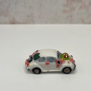 May include: A miniature white ceramic car decorated with pink and green floral accents. The small vehicle has black wheels and a glossy finish, suggesting a decorative or collectible item.