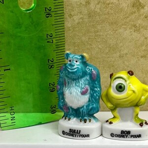 May include: Two ceramic figurines of characters from the movie Monsters, Inc. The blue Sully figure is next to a yellow Bob figure. A green ruler shows the figurines are about 3 inches tall. The bases read "SULLI" and "BOB" with the Disney/Pixar logo.