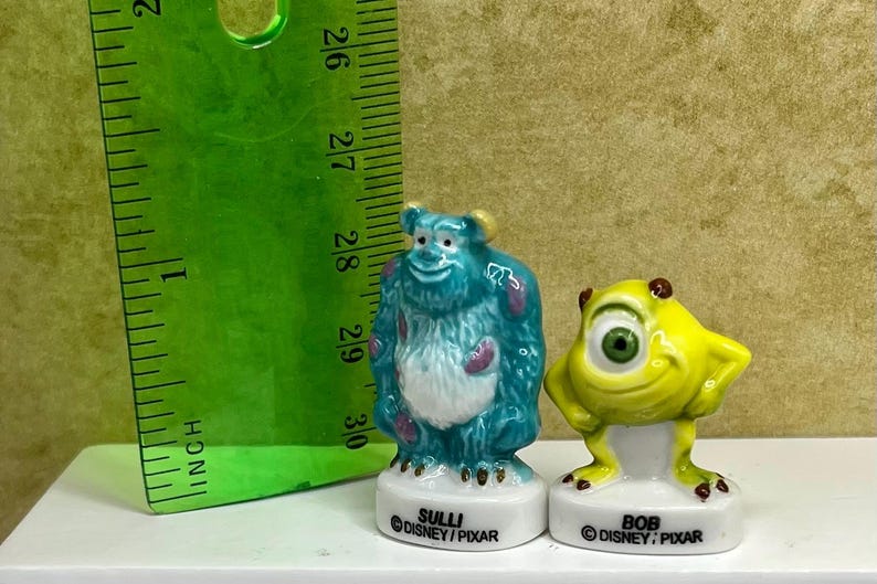 May include: Two ceramic figurines of characters from the movie Monsters, Inc. The blue Sully figure is next to a yellow Bob figure. A green ruler shows the figurines are about 3 inches tall. The bases read "SULLI" and "BOB" with the Disney/Pixar logo.