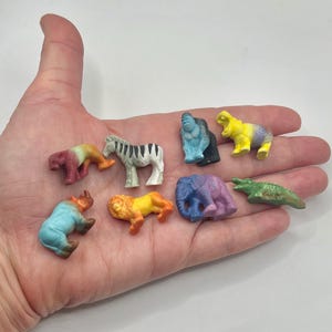 May include: A collection of small, colorful animal figurines, including a zebra, lion, elephant, and crocodile. The figurines are painted in various colors and appear to be made of plastic or a similar material. The animals are displayed on a light background.