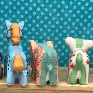 May include: A collection of colorful ceramic animal figurines, including dogs, giraffes, and ducks, arranged on a wooden shelf. The figurines feature various patterns and colors, such as blue, yellow, and pink, and are set against a teal background with white polka dots.