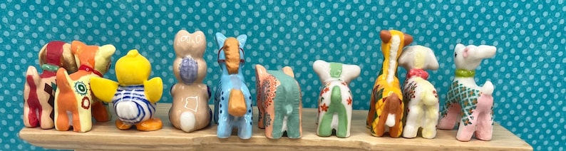 May include: A collection of colorful ceramic animal figurines, including dogs, giraffes, and ducks, arranged on a wooden shelf. The figurines feature various patterns and colors, such as blue, yellow, and pink, and are set against a teal background with white polka dots.