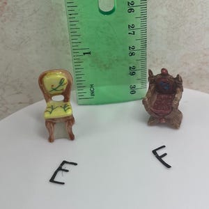 May include: Miniature dollhouse furniture set featuring two small chairs. One chair has a yellow seat and back with floral details, the other is a brown armchair. A green ruler is next to the chairs. The chairs are approximately 1 inch in height.