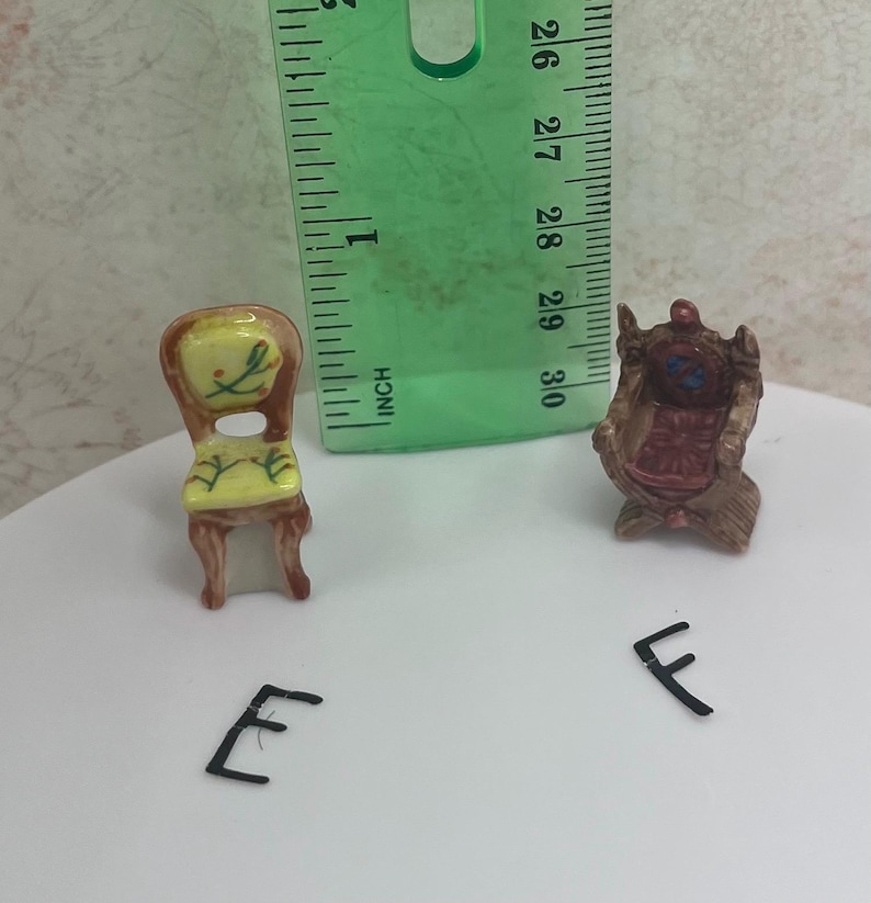 May include: Miniature dollhouse furniture set featuring two small chairs. One chair has a yellow seat and back with floral details, the other is a brown armchair. A green ruler is next to the chairs. The chairs are approximately 1 inch in height.