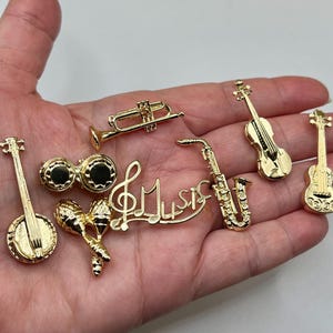 May include: A collection of gold-colored miniature musical instruments and a "Music" wordmark. Instruments include a trumpet, saxophone, violin, banjo, maracas, bongo drums, and a guitar. The items are small and detailed.