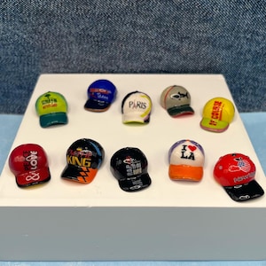 May include: A collection of miniature baseball caps in various colors, displayed on a white surface. Each cap features unique designs, including text and graphics. The caps are in red, black, yellow, green, blue, and white.