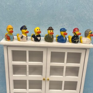 May include: A collection of seven yellow rubber duck figurines, each dressed in a different outfit, including a construction worker, a sailor, and a chef. The ducks are displayed on top of a white cabinet with glass doors, against a light blue background.