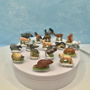 May include: A collection of small, ceramic animal figurines, including a badger, fox, and squirrel. Each figurine is painted with realistic details and mounted on a small, white base with French text. The figurines are displayed on a white rotating platform.
