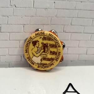 May include: A small, round tambourine with a yellow face featuring a classical illustration and the words "MUSIQUE - MUSE". The tambourine has silver jingles and an orange rim. A black letter "A" is at the bottom.