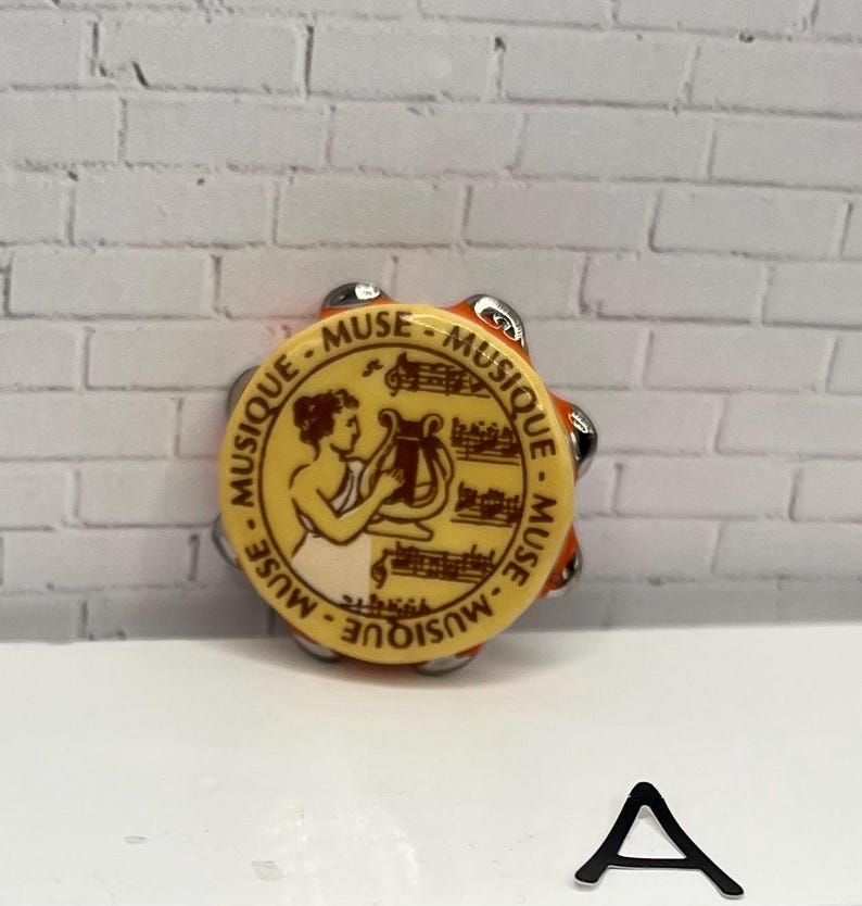 May include: A small, round tambourine with a yellow face featuring a classical illustration and the words "MUSIQUE - MUSE". The tambourine has silver jingles and an orange rim. A black letter "A" is at the bottom.