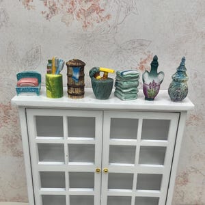 May include: Miniature dollhouse bathroom accessories, including a blue chair with a pink cushion, a green container with brushes, a woven basket, a green container with a yellow faucet, folded towels, and decorative bottles. The items are arranged on a white cabinet.