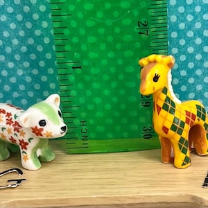 May include: Two miniature ceramic figurines: a bear with floral patterns and a giraffe with a diamond pattern. The giraffe is approximately 2 inches tall. The figurines are on a wooden surface with a green ruler in the background.