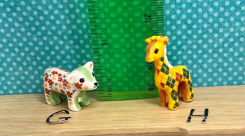 May include: Two miniature ceramic figurines: a bear with floral patterns and a giraffe with a diamond pattern. The giraffe is approximately 2 inches tall. The figurines are on a wooden surface with a green ruler in the background.
