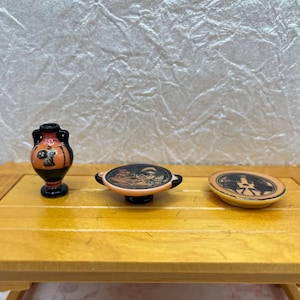 May include: Miniature ceramic Greek pottery set, including a vase, a plate, and a serving dish. The pieces are painted with black and orange figures on a cream background. The set is displayed on a small wooden table.