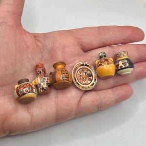 May include: A collection of six miniature ceramic items including four small vases, one bottle, and one plate. Each piece features intricate geometric and figurative patterns in shades of orange, yellow, black, white, and brown, resting on a palm.