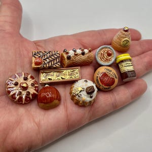 May include: A collection of miniature ceramic food items, including cakes, pastries, and a jar of preserves. The items are in various shapes and colors, with intricate details. The collection includes a small round item with the text "CHAUD CHAUD".