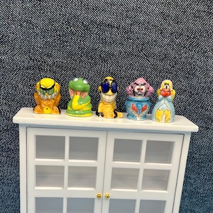 May include: Five colorful, cartoonish figurines are displayed on a white cabinet. The figurines include a yellow monster, a green snake, a yellow character with sunglasses, a purple-haired character in a blue vase, and a blonde character in a blue dress.